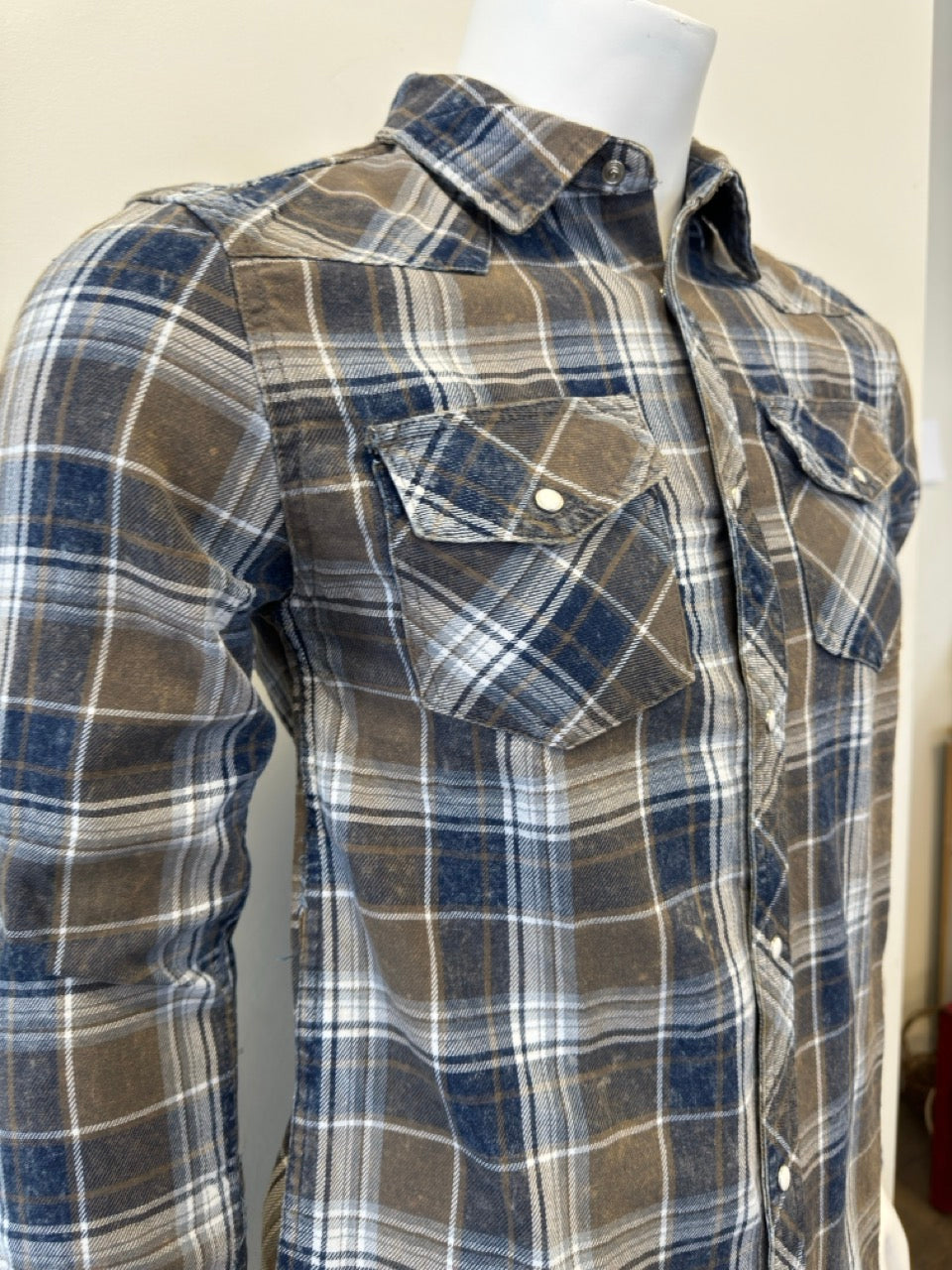 A mannequin wears the Shirt Long Sleeve - OLIVE by Silver Jeans Co., showcasing a plaid design in shades of brown, blue, and white. Crafted from 100% cotton, this button-up shirt features two front chest pockets with buttoned flaps. The mannequin's hand is partially visible on the right side as it stands against a plain, light-colored background.