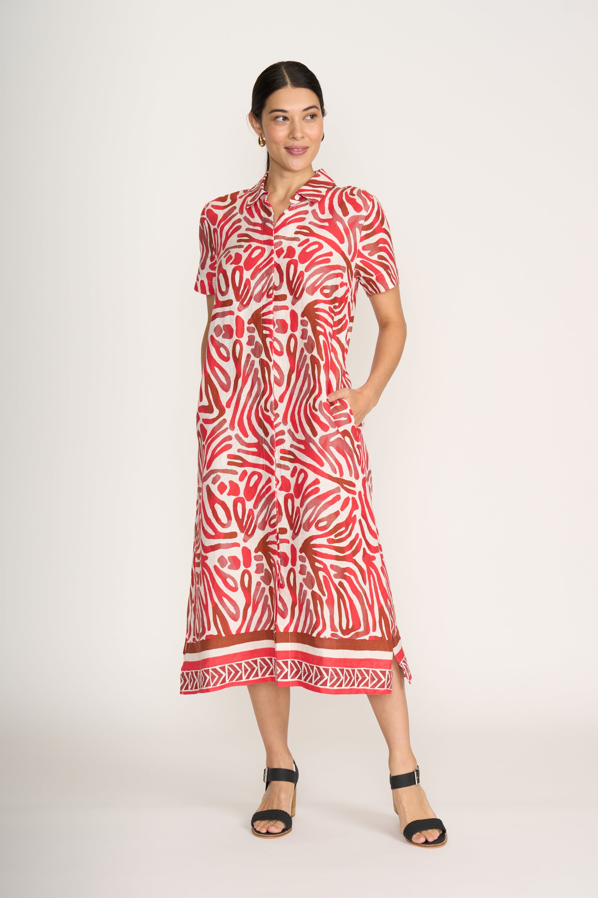 A woman smiles and looks to the side wearing the FOIL Advanced Interaction Dress Prowl FO8296, a red and white prowl print linen midi dress with short sleeves, button-down front, and pockets. She pairs it with black sandals and has her hair pulled back.