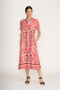 A woman smiles and looks to the side wearing the FOIL Advanced Interaction Dress Prowl FO8296, a red and white prowl print linen midi dress with short sleeves, button-down front, and pockets. She pairs it with black sandals and has her hair pulled back.