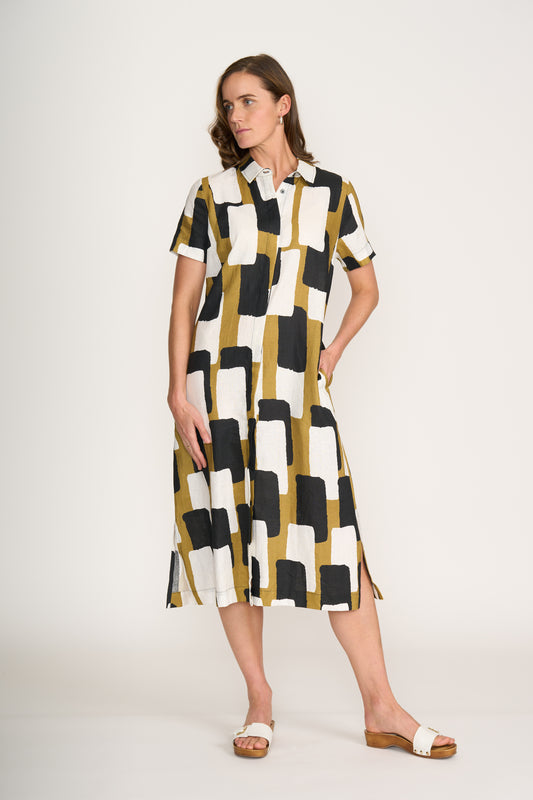 A woman stands against a plain background wearing the FOIL Advanced Interaction Dress Art Box FO8296, a short-sleeve, button-up linen dress featuring a bold black, white, and mustard yellow print, styled with white sandals.
