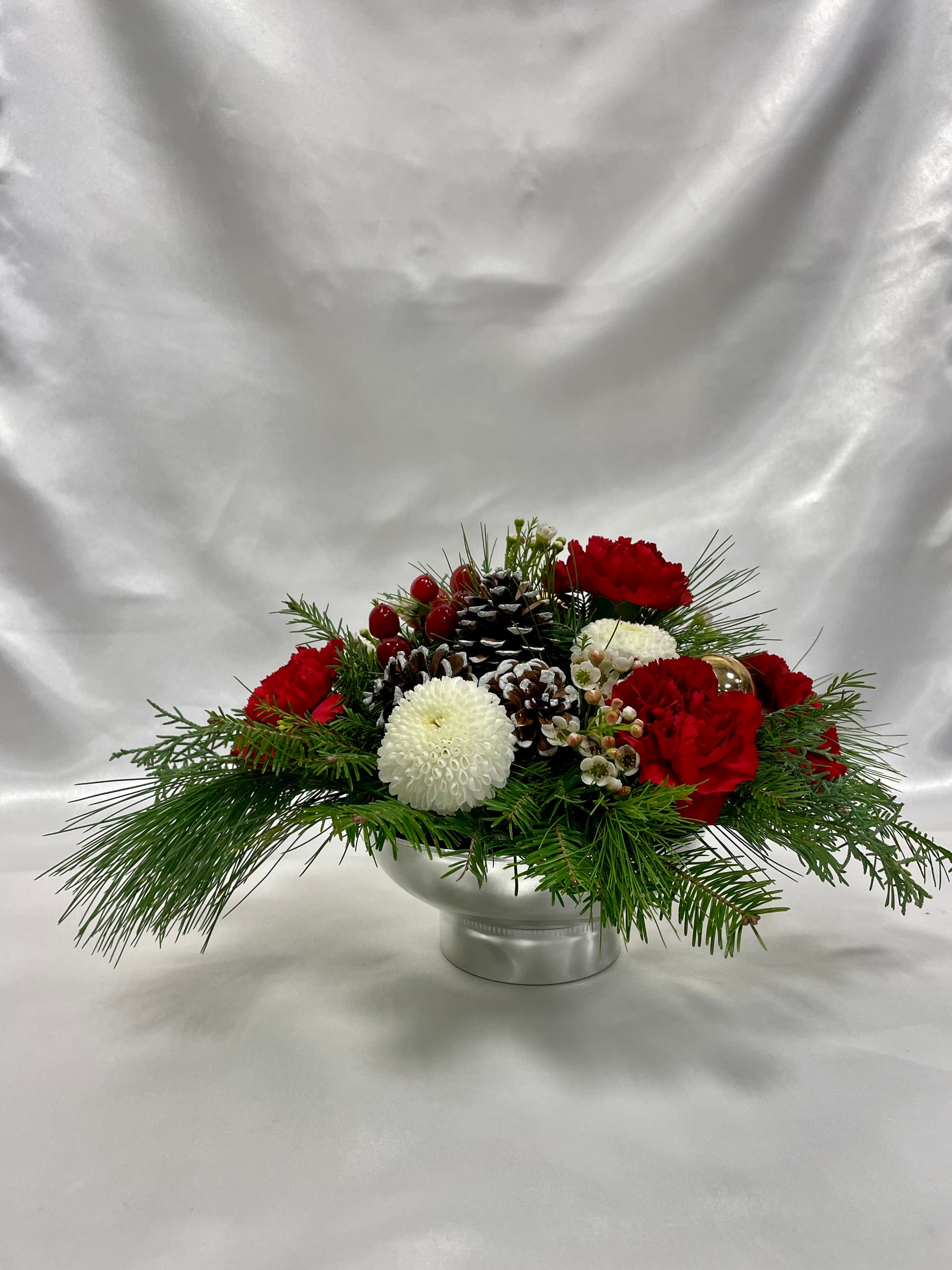 The "Simple Christmas" arrangement by Posies Flowers & Fashion, featuring red and white blooms, pinecones, and lush greenery in a silver vase, effortlessly infuses any setting with festive joy against a white fabric background.