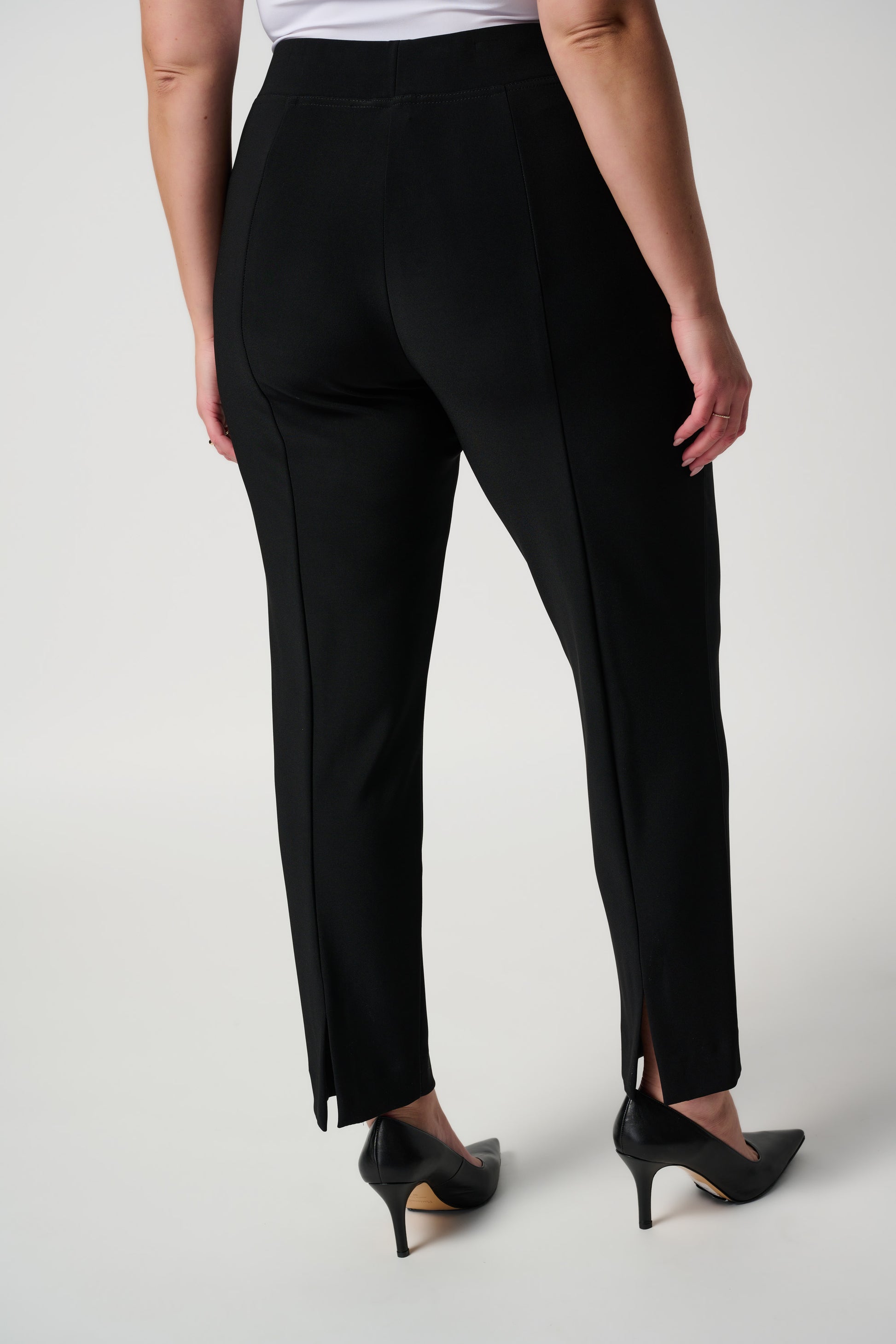 A person wearing the Classic Straight Pant in black by Joseph Ribkoff with slits at the ankles and black pointed heels, standing with their back to the camera. The person is also wearing a white top made of silky knit material. The background is plain white.