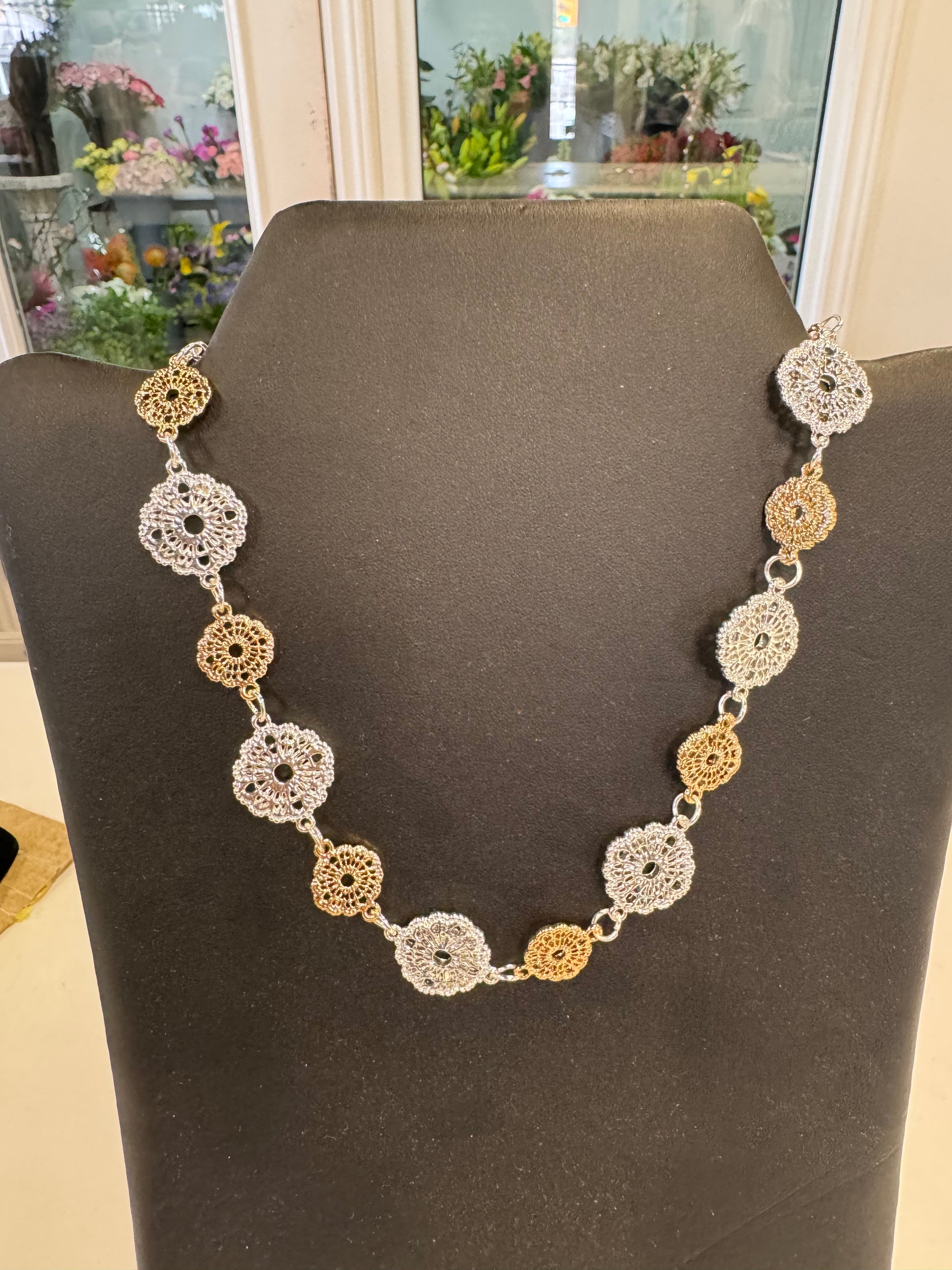 A stunning piece of jewelry from Posies Flowers & Fashion Inc., the Bronzes and Silver Coloured Necklace, is displayed on a black stand, featuring an alternating pattern of silver and gold circular filigree medallions linked together. The backdrop includes reflections of flowers in a floral shop, highlighting its unique design.