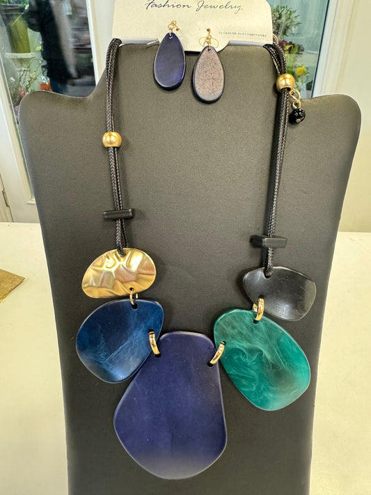 A black display stand showcases the timeless Blue, Black, and Gold Necklace with Earrings set by Posies Flowers & Fashion Inc. The statement necklace features large abstract pendants in gold, black, deep blue, and teal on a black cord with gold beads. Above it, matching blue and black teardrop-shaped earrings are pinned.