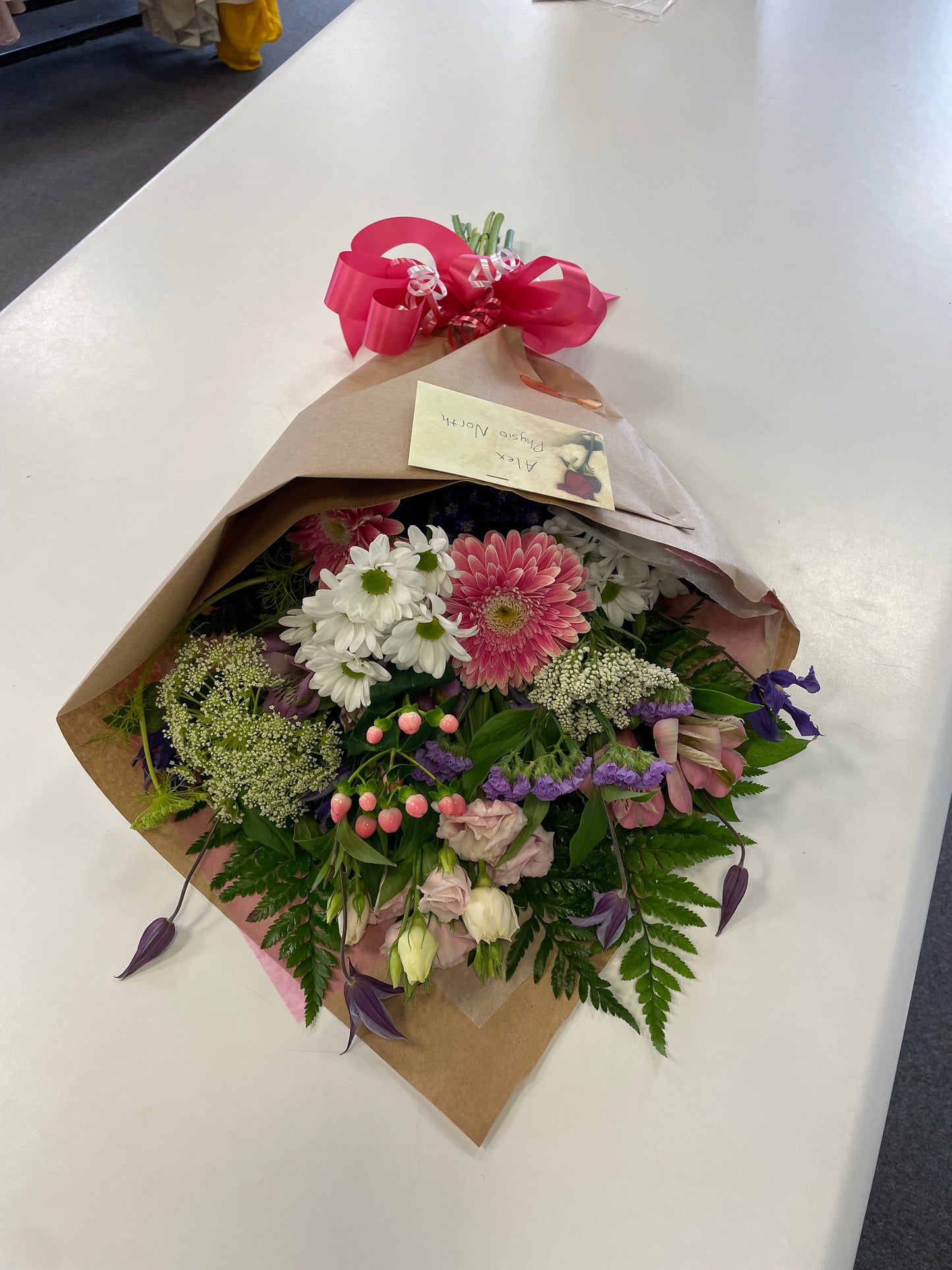 A stunning bouquet called Designer’s Choice Freshcuts from Posies Flowers & Fashion is resting on a white surface. The arrangement features vibrant orange and pink roses, cheerful yellow chrysanthemums, elegant white flowers, and assorted green foliage. It is partially wrapped in brown paper and elegantly tied with a burgundy ribbon.