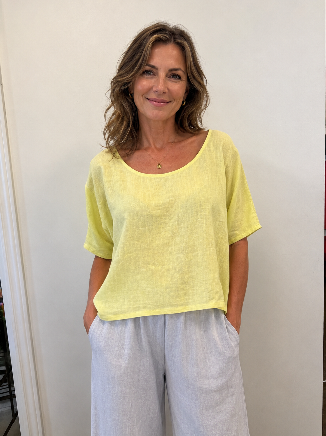 A woman with wavy brown hair smiles in front of an off-white wall, wearing the ETERNELLE Top Yellow 6-SS26125-9—a loose Italian-made linen women's top—and light gray pants, with her hands in her pockets.