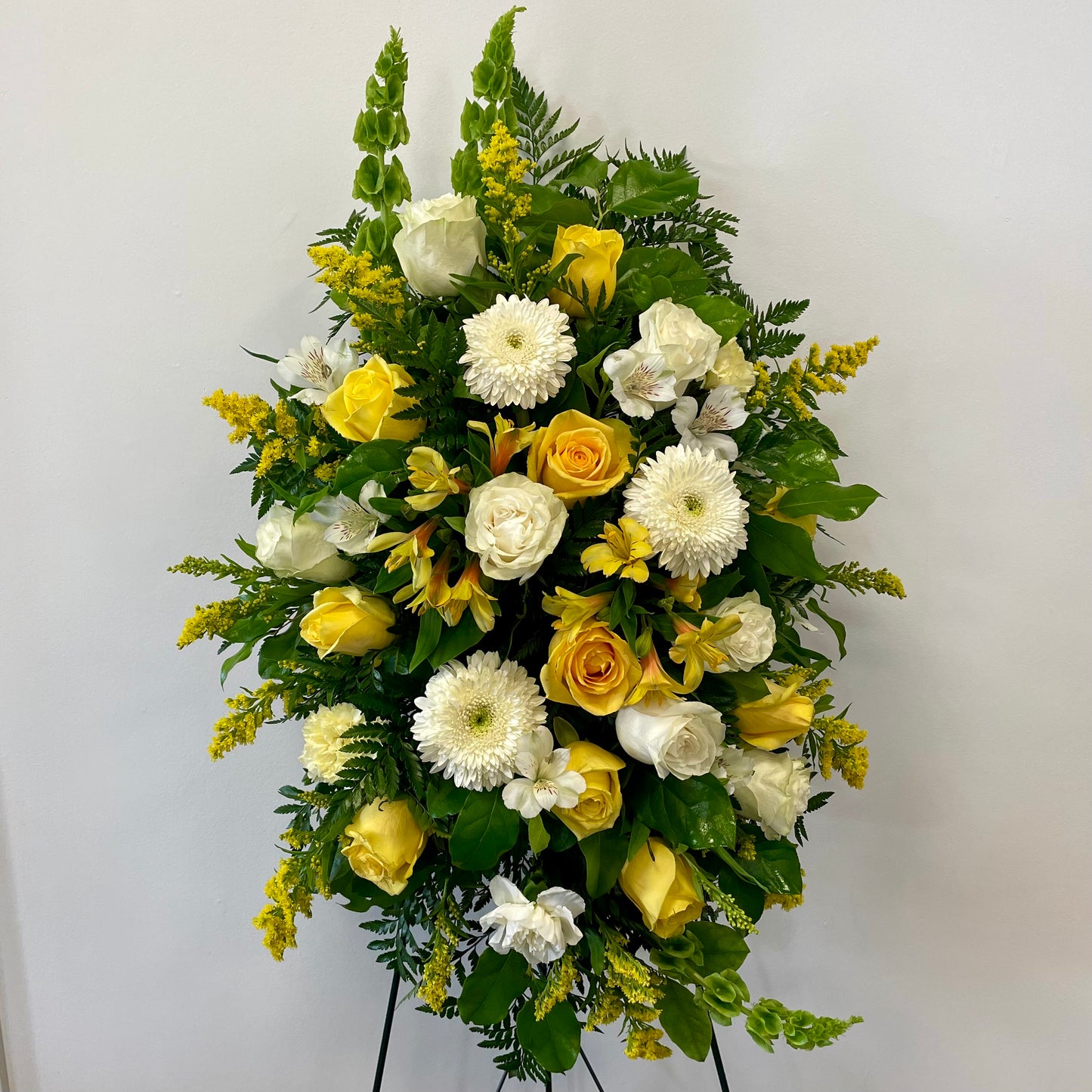 The Sunlit Serenity arrangement from Posies Flowers & Fashion showcases a striking mix of yellow roses, white roses, white chrysanthemums, and various other yellow and white blossoms, all artfully set against a white wall. Accentuated with lush green leaves and ferns, this elegant tribute creates a vibrant display reminiscent of a funeral hanging spray.