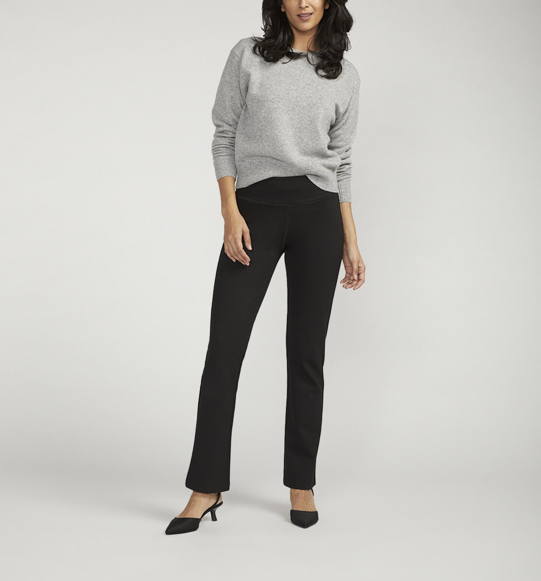 A person is posing against a plain background, wearing a light gray long-sleeve sweater paired with JAG's Mid Rise Bootcut Pull-On Pants. They have one hand on their hip and are sporting black pointed-toe heeled shoes. Their dark hair flows loose and wavy.