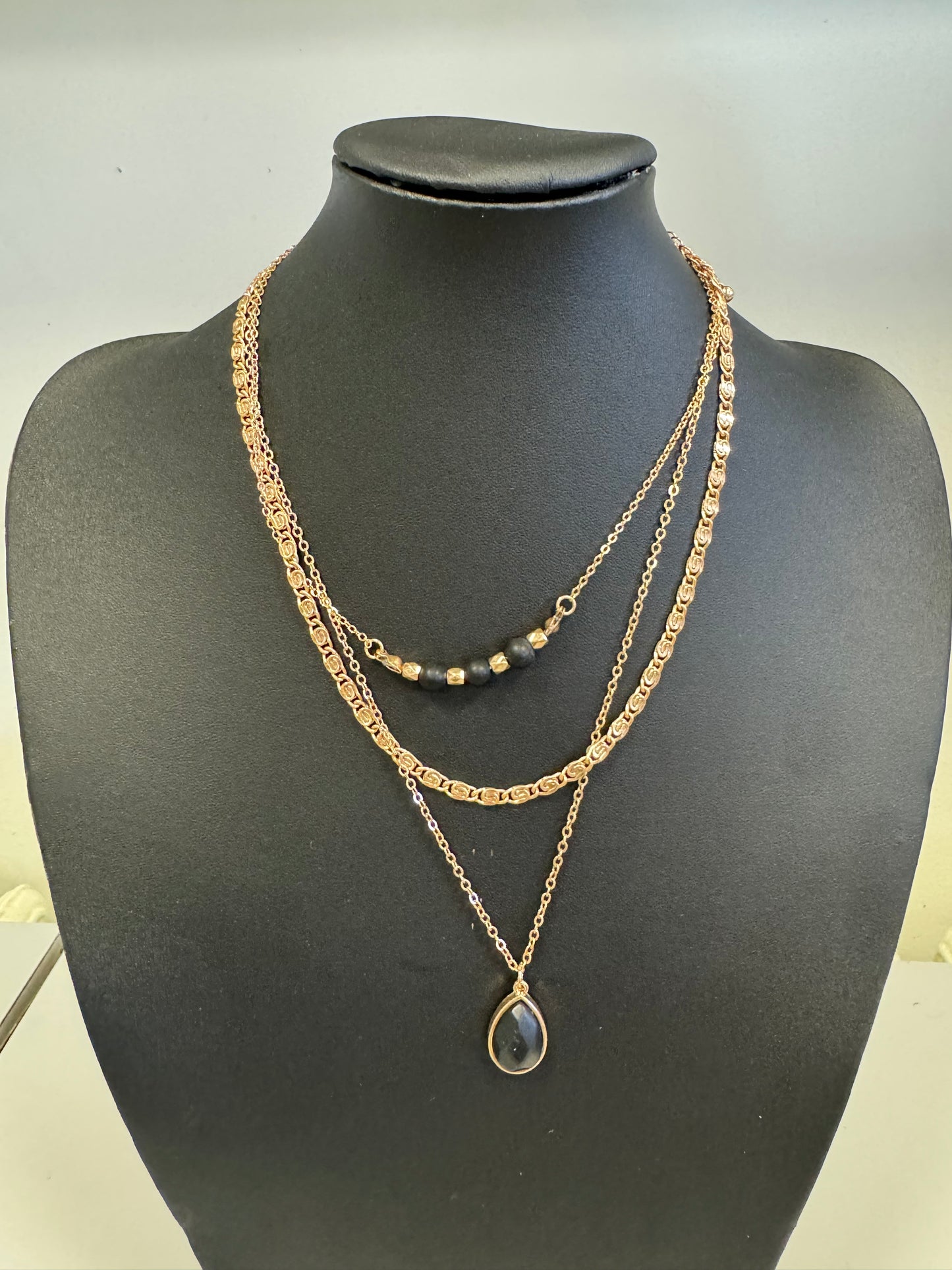 The Hanna 22 Necklace from Elise Accessories is elegantly displayed on a black bust. This three-layered chain necklace features the top two layers with gold chains, while the shortest is adorned with small black beads. The longest layer showcases a teardrop-shaped pendant with a dark stone, making it a versatile addition to any jewelry collection.