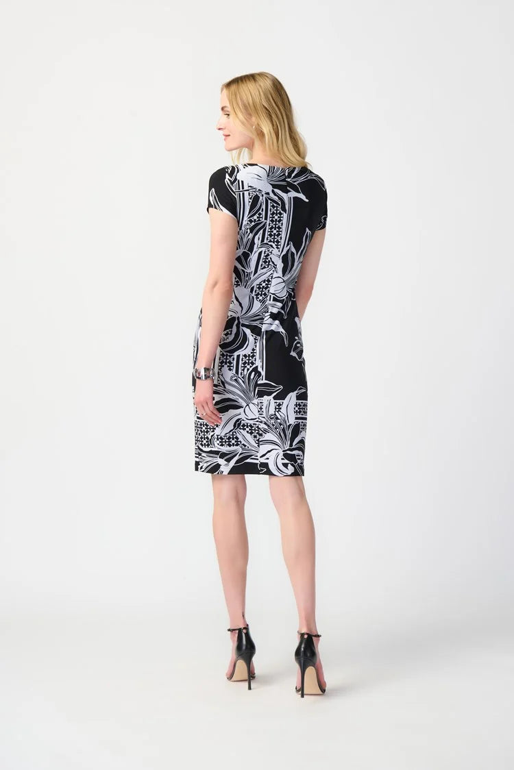 A woman with long blonde hair stands against a plain background, wearing the Joseph Ribkoff Floral Print Silky Knit Sheath Dress 241284. This short-sleeve black dress features a chic silhouette with large white flowers and geometric accents along the border. She has a neutral expression and one hand placed on her hip.