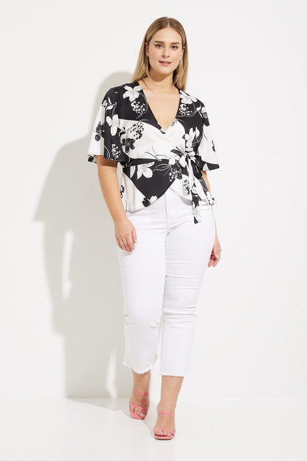 A woman with straight blonde hair exudes casual elegance, wearing a Vanilla/Black Wrap Front Top Style 232058 by Joseph Ribkoff, paired with white pants. She stands against a plain white background, one hand casually resting on her hip, looking confidently at the camera.