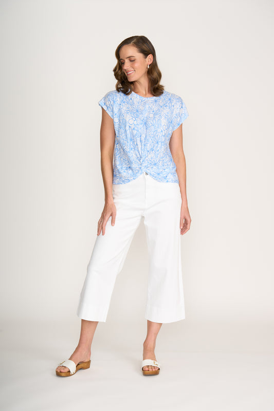 A woman with shoulder-length brown hair smiles slightly while looking left, wearing the FOIL Do The Twist Top FO8258 in Calypso and white wide-leg pants against a plain white background, paired with sandals.