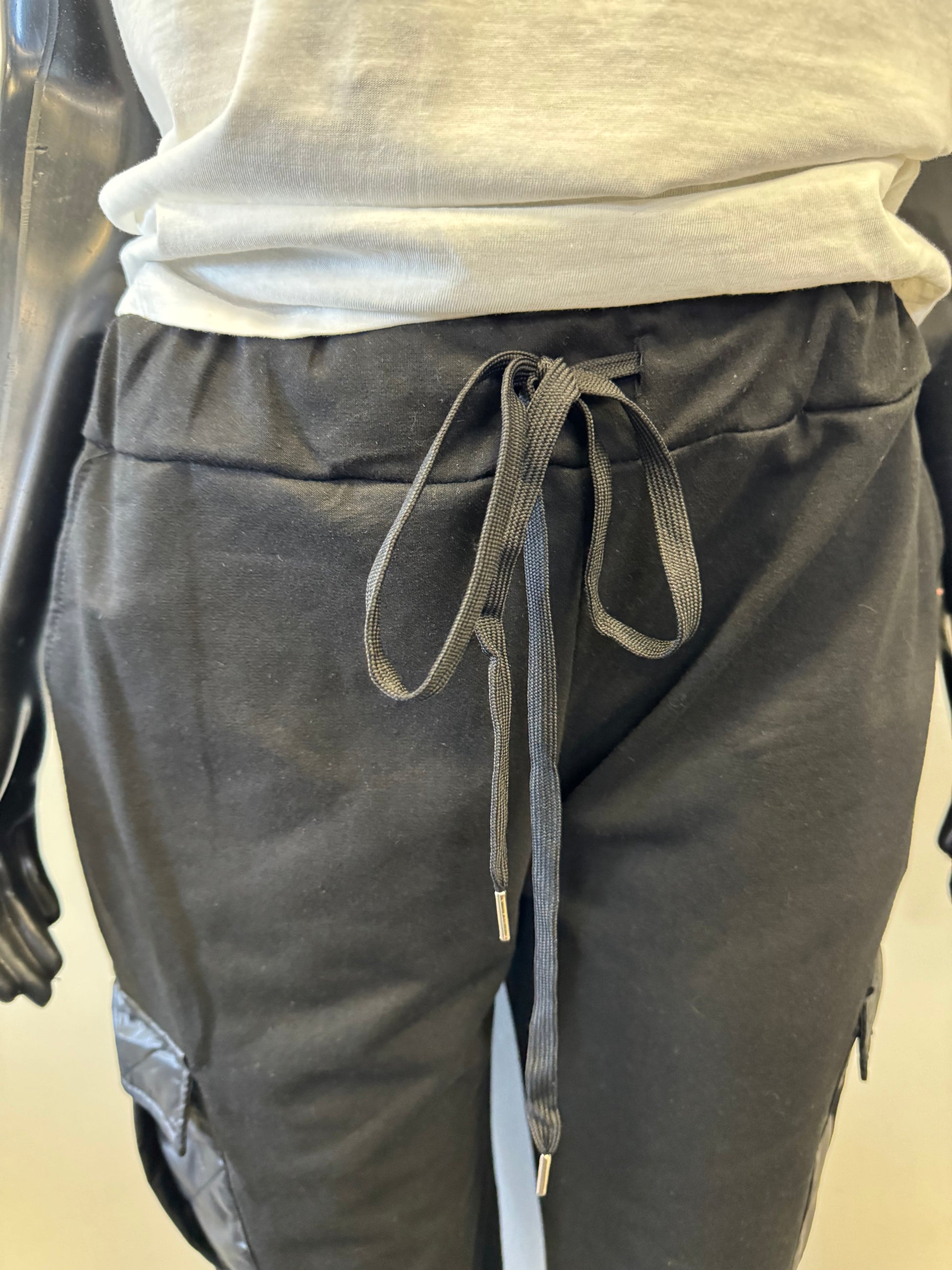 A mannequin dressed in ETERNELLE's Black Joggers with patch pockets, featuring a drawstring waist and elastic cuffs. The jogger pants, made in Italy, have stylish cargo pockets with a glossy zippered detail and are paired with a plain white top. The mannequin's hands are visible, resting by its sides.