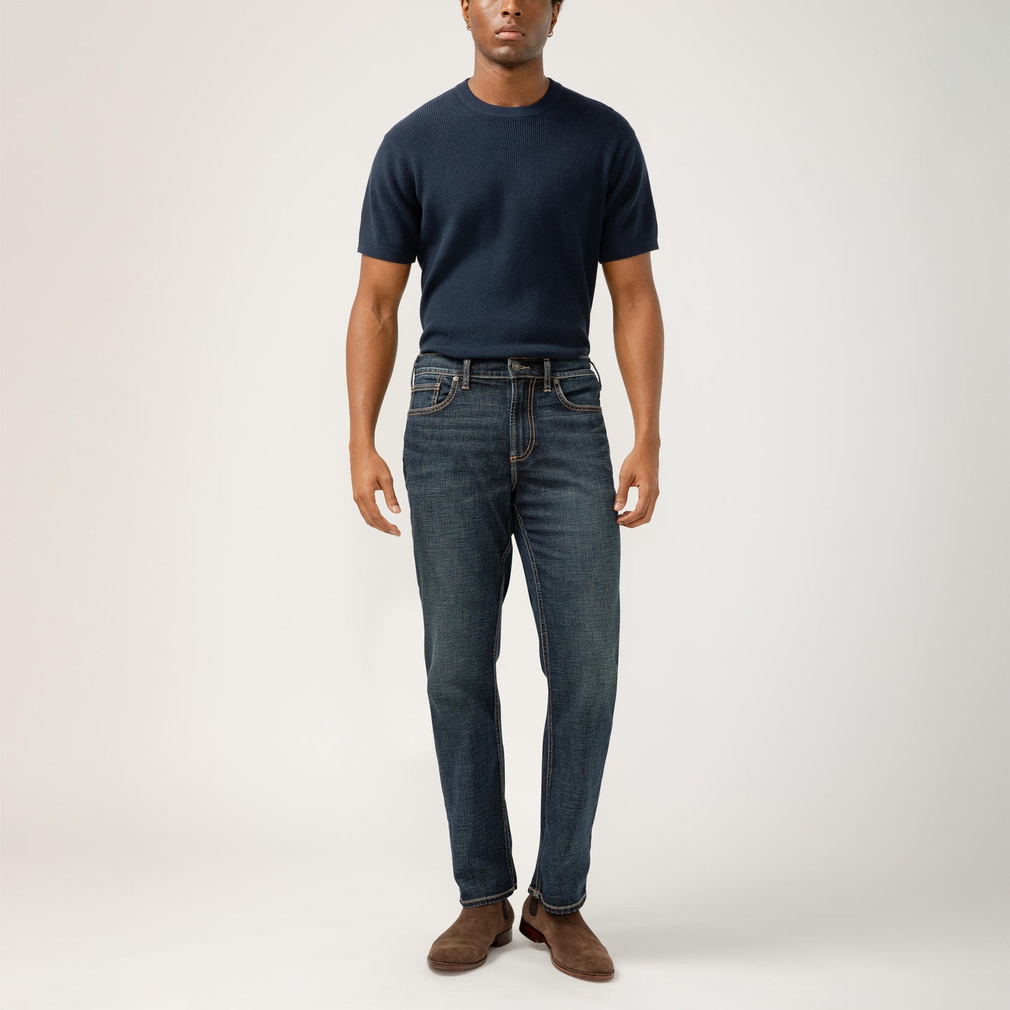 A person stands against a plain background wearing a navy blue, classic fit, short-sleeve shirt tucked into Grayson Classic Fit Straight Leg Silver Jeans M23425EFS409 by Silver Jeans Co. with brown suede shoes. Their arms rest comfortably by their sides, and their shoes contrast with the white backdrop. Style code: SIL-M23425EFS409-IND.