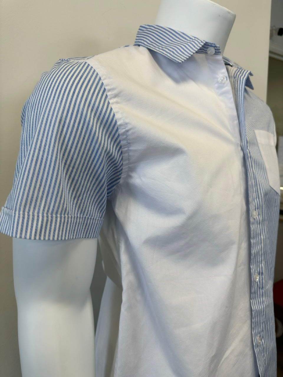 A mannequin showcases the Men’s Cotton Shirt - STRIPE WHITE/BLUE by Silver Jeans Co., which features a button-up style with alternating vertical white and blue stripes on the right side and pocket. The left side and sleeves are solid white, prominently displayed against a plain backdrop that accentuates the garment's unique design.