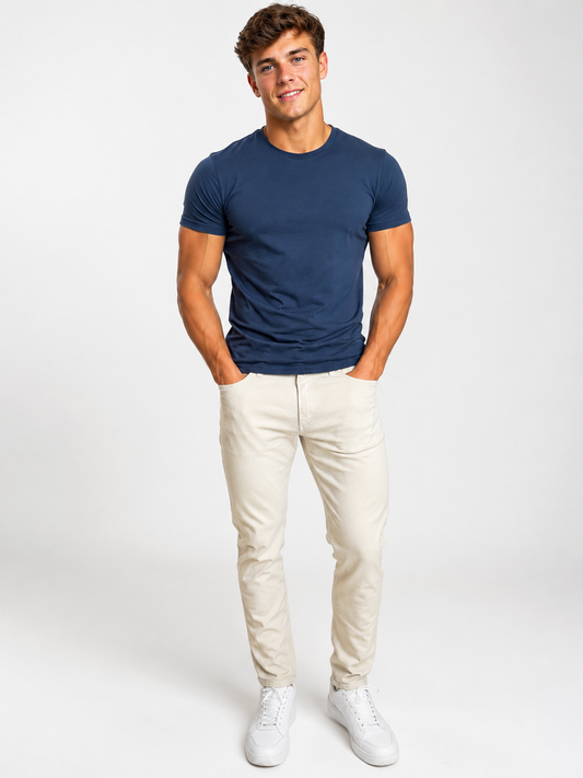 A young man smiles with hands in pockets, wearing a fitted navy t-shirt, Silver Jeans Co. Machray Classic Fit Slim Straight Leg Jeans in Stone (M63410LBS659), and white sneakers against a plain white background.