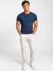 A young man smiles with hands in pockets, wearing a fitted navy t-shirt, Silver Jeans Co. Machray Classic Fit Slim Straight Leg Jeans in Stone (M63410LBS659), and white sneakers against a plain white background.
