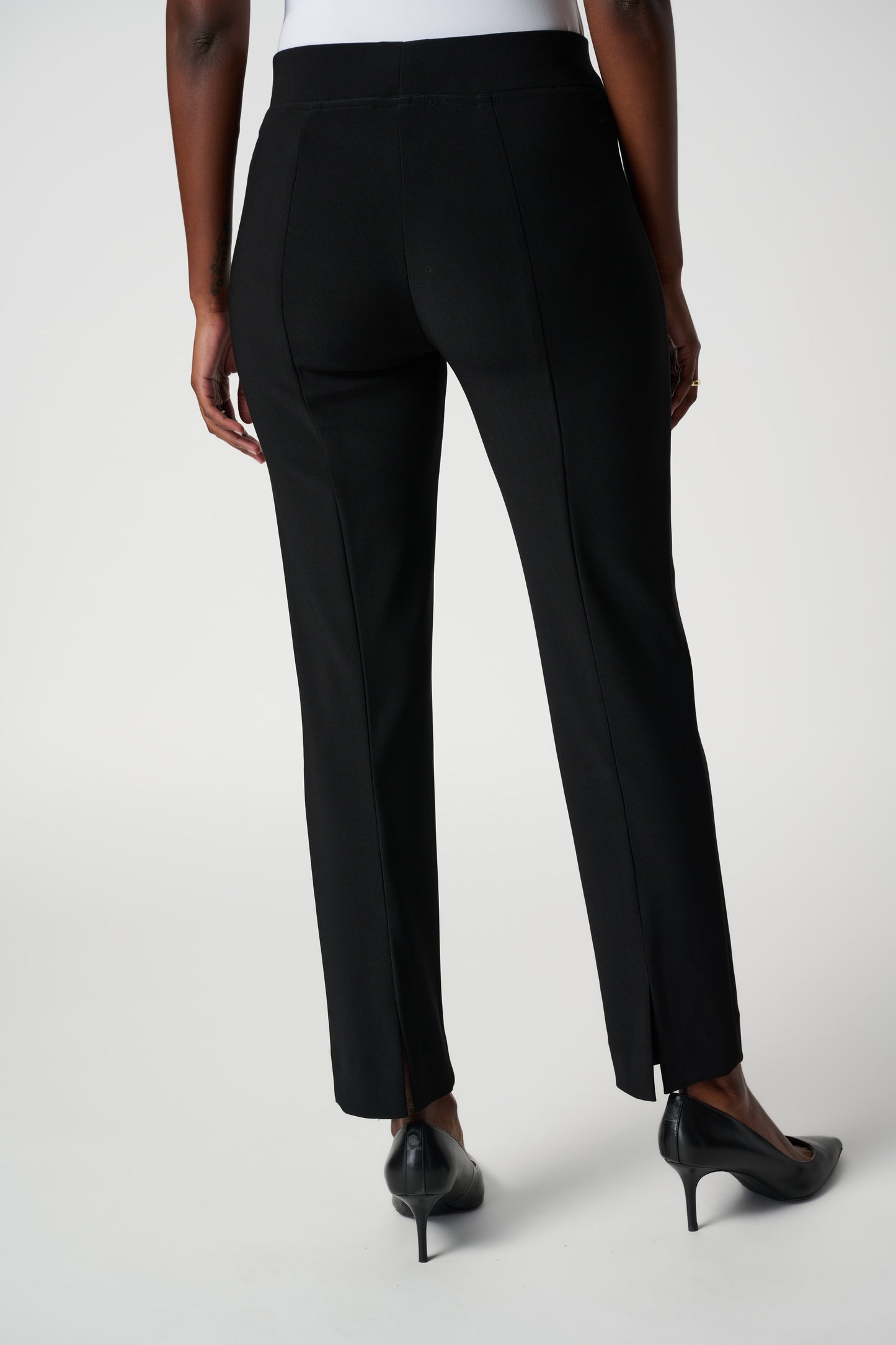 A person wearing the Classic Straight Pant in black by Joseph Ribkoff with slits at the ankles and black pointed heels, standing with their back to the camera. The person is also wearing a white top made of silky knit material. The background is plain white.