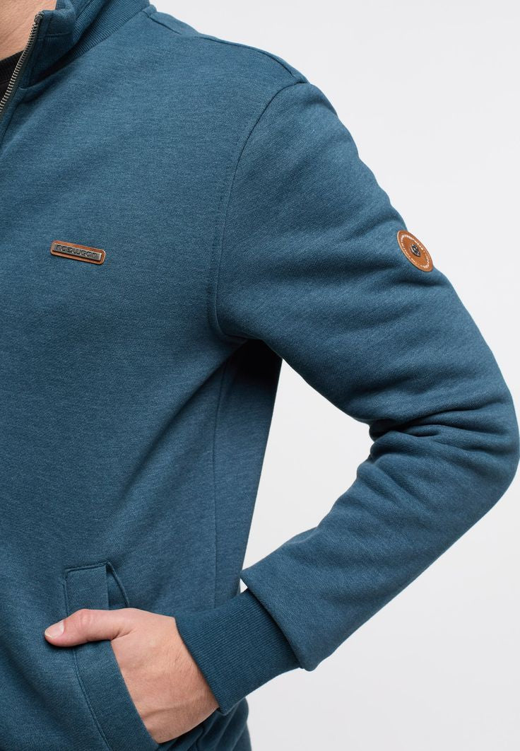 A person wearing the Ragwear Sweatshirt Zip - TRAYNE TURQUOISE by RAGWEAR is shown from the shoulders to the mid-thigh. Their right hand is visible, resting by their side. The turquoise sweatshirt features a small brown patch with writing near the bottom hem, just above their dark-colored pants.