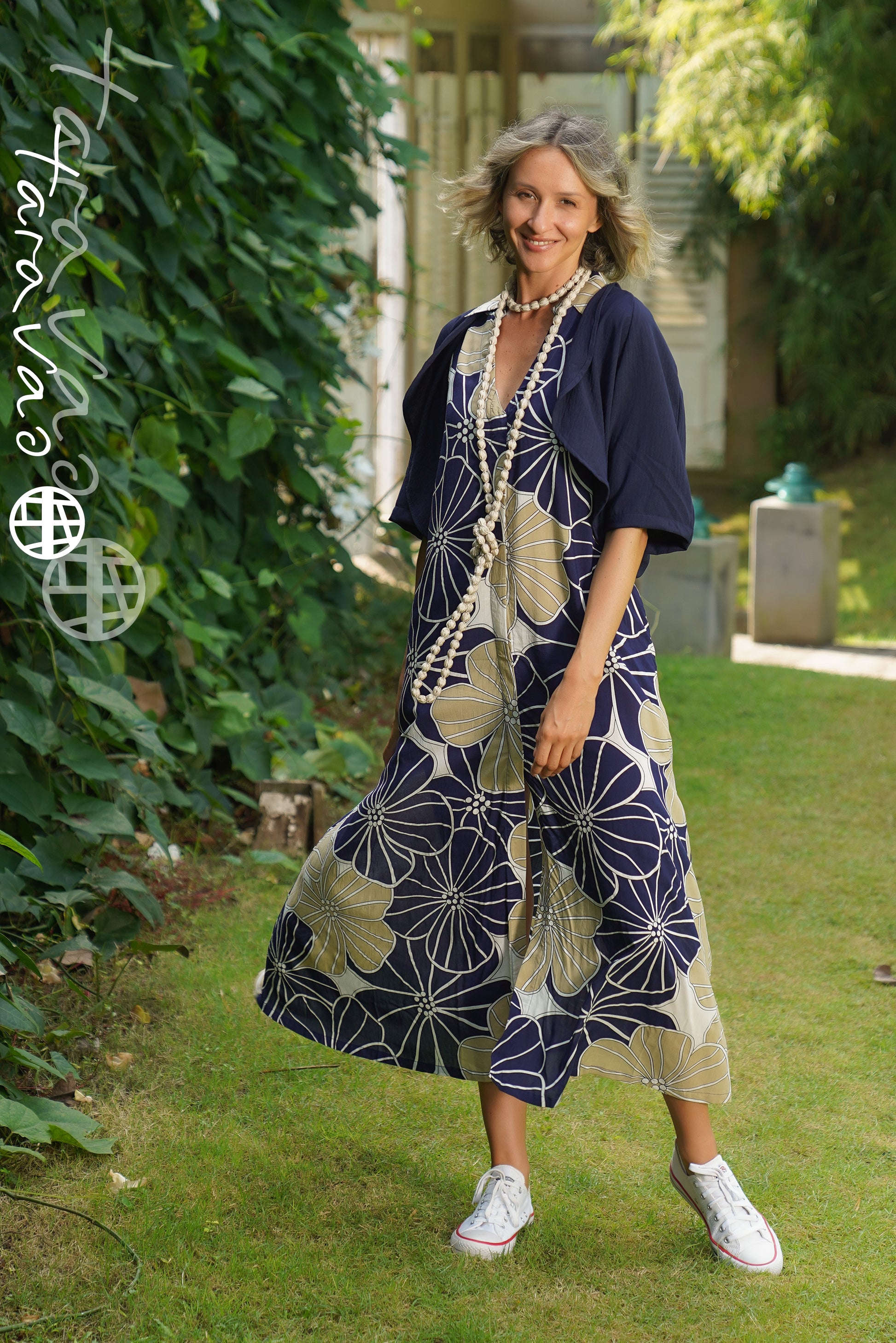 A smiling woman stands in a sunlit garden wearing the TaraVao Dress Marilou 9048 N, paired with a navy shrug, long beaded necklaces, and white sneakers amid lush green plants.