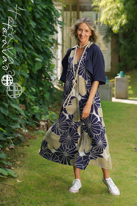 A smiling woman stands in a sunlit garden wearing the TaraVao Dress Marilou 9048 N, paired with a navy shrug, long beaded necklaces, and white sneakers amid lush green plants.