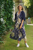 A smiling woman stands in a sunlit garden wearing the TaraVao Dress Marilou 9048 N, paired with a navy shrug, long beaded necklaces, and white sneakers amid lush green plants.