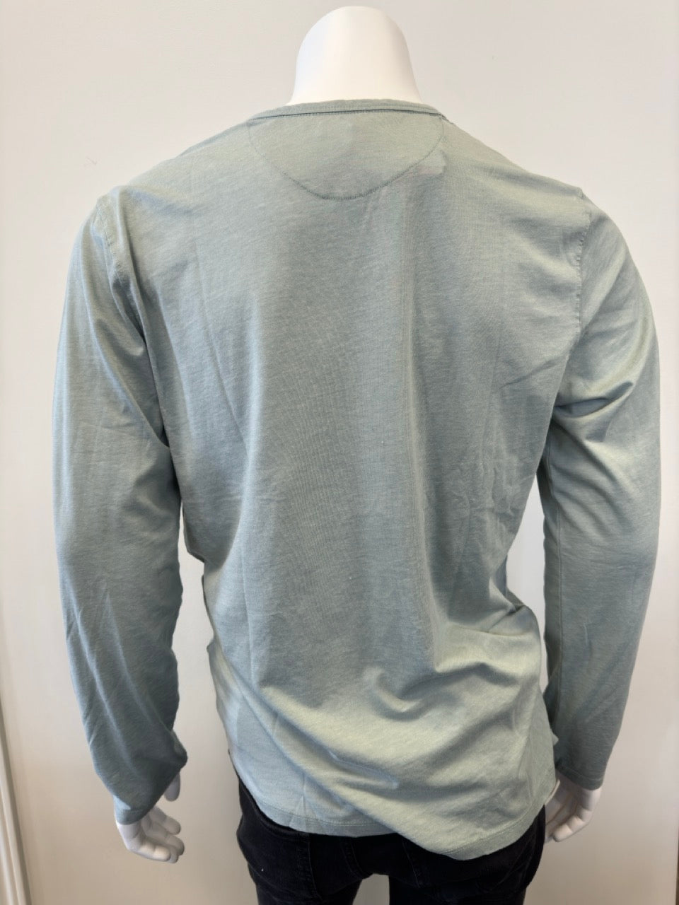 A mannequin dressed in the Silver Jeans Co. Shirt Long Sleeve - SAGE, featuring a classic 3-button design along the V-neckline. The sage green shirt, made from a comfortable cotton-polyester blend, is paired with black pants. The background is plain and white.