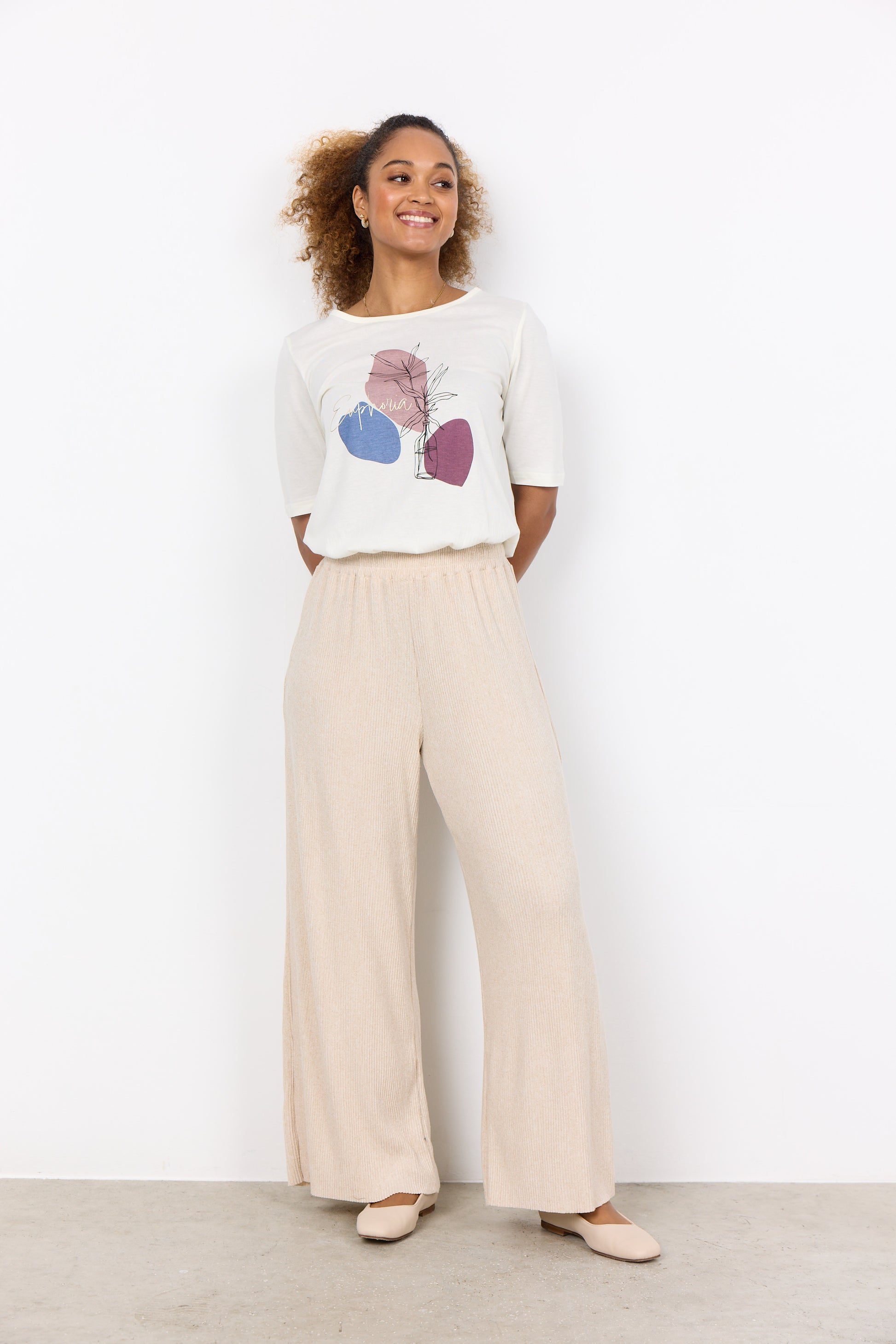 A woman with curly hair pulled back, wearing the stylish T-Shirt Felicity Woodrose SC-FELECITY FP 475 from RAGWEAR featuring abstract shapes and the word "Euphoria," stands in front of a white background. She pairs it with light-colored pants, making her look effortlessly chic.