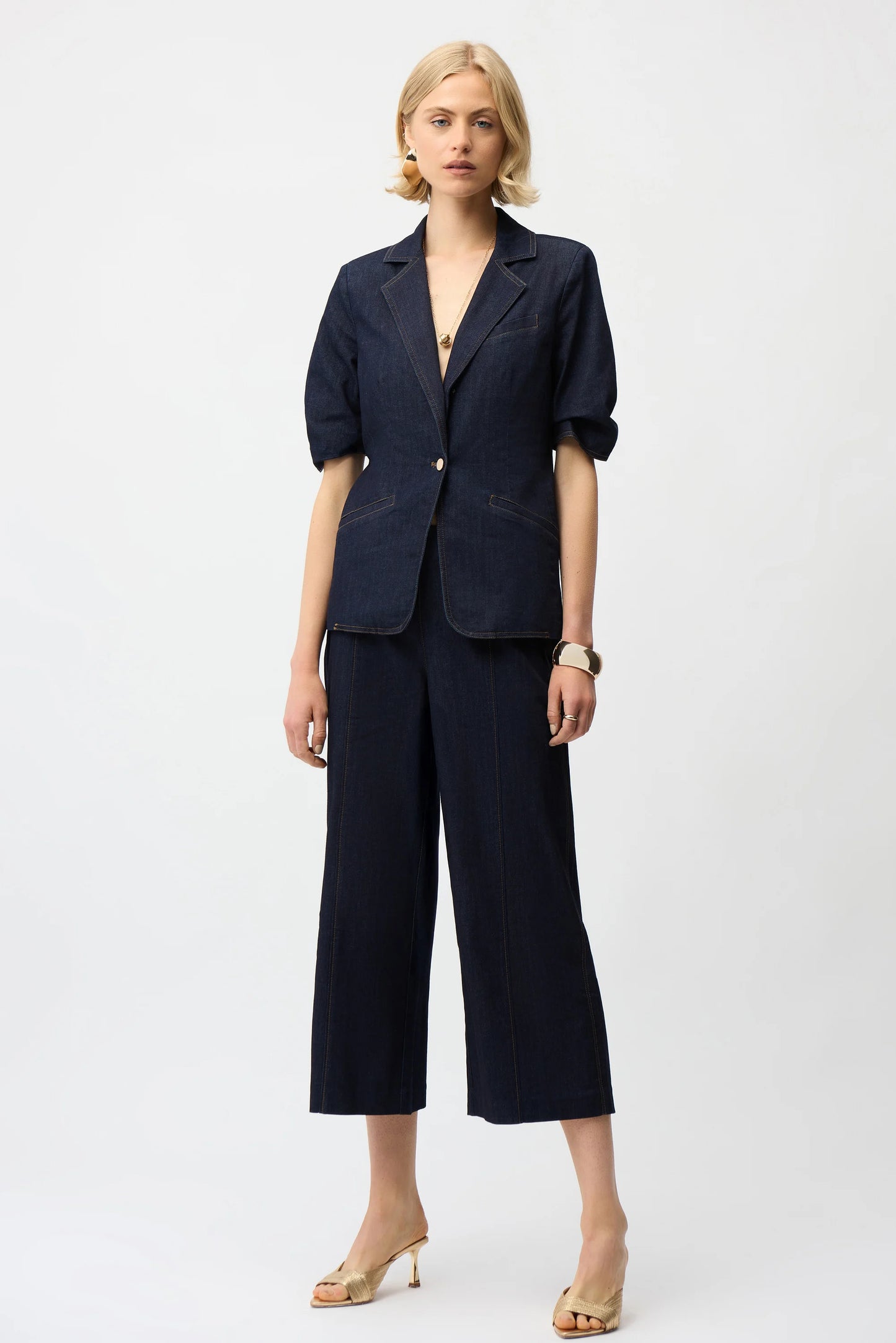 A woman poses against a plain background in Joseph Ribkoff’s Culotte Jeans With Exposed Side Zipper 261955, styled with a modern denim blazer, gold heels, and simple jewelry.