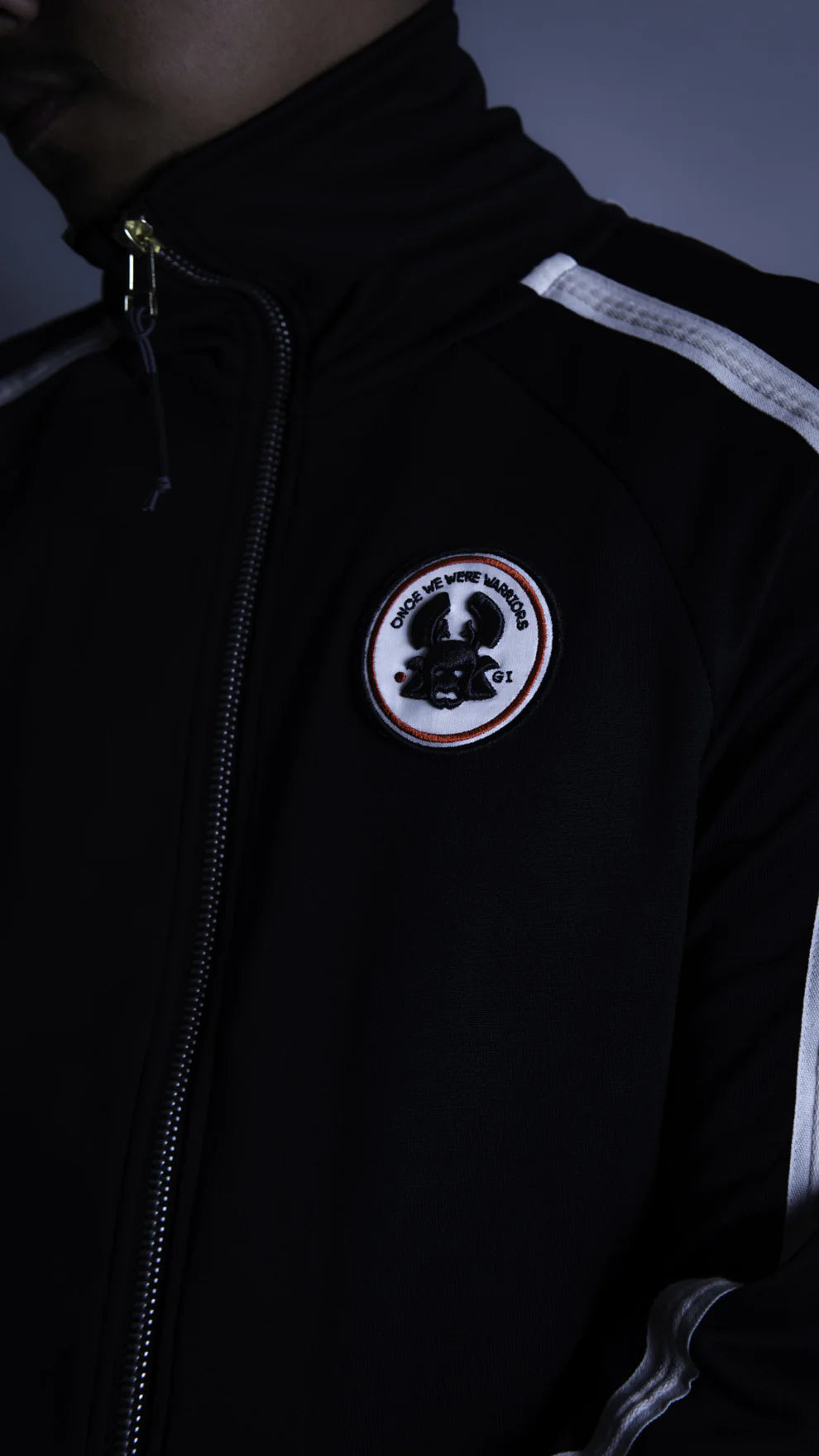 A person with short, curly hair stands in a dark, minimalist studio wearing the Once We Were Warriors Haku Track Jacket - Black and matching pants, which are both adorned with white stripes down the sleeves and legs. The sportswear set features a circular emblem on the chest and thigh. They are also wearing black sneakers and looking downward.