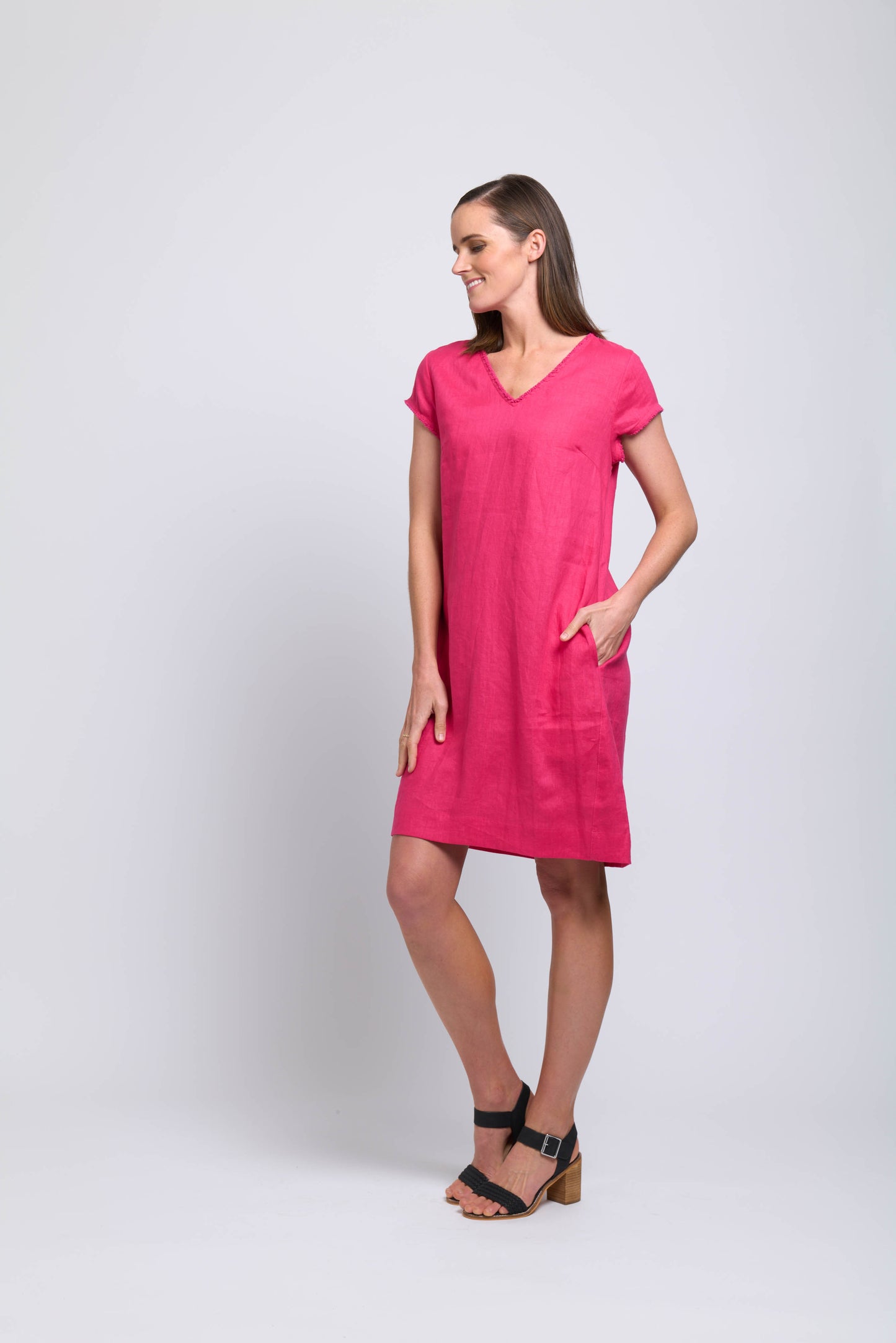 A woman stands against a plain light background, smiling in the FOIL Fringe Festival Dress FO7907 Pink Flash—a bright pink, knee-length dress with short sleeves and a V-neckline. She has her left hand in her pocket and looks to the side wearing black heeled sandals.