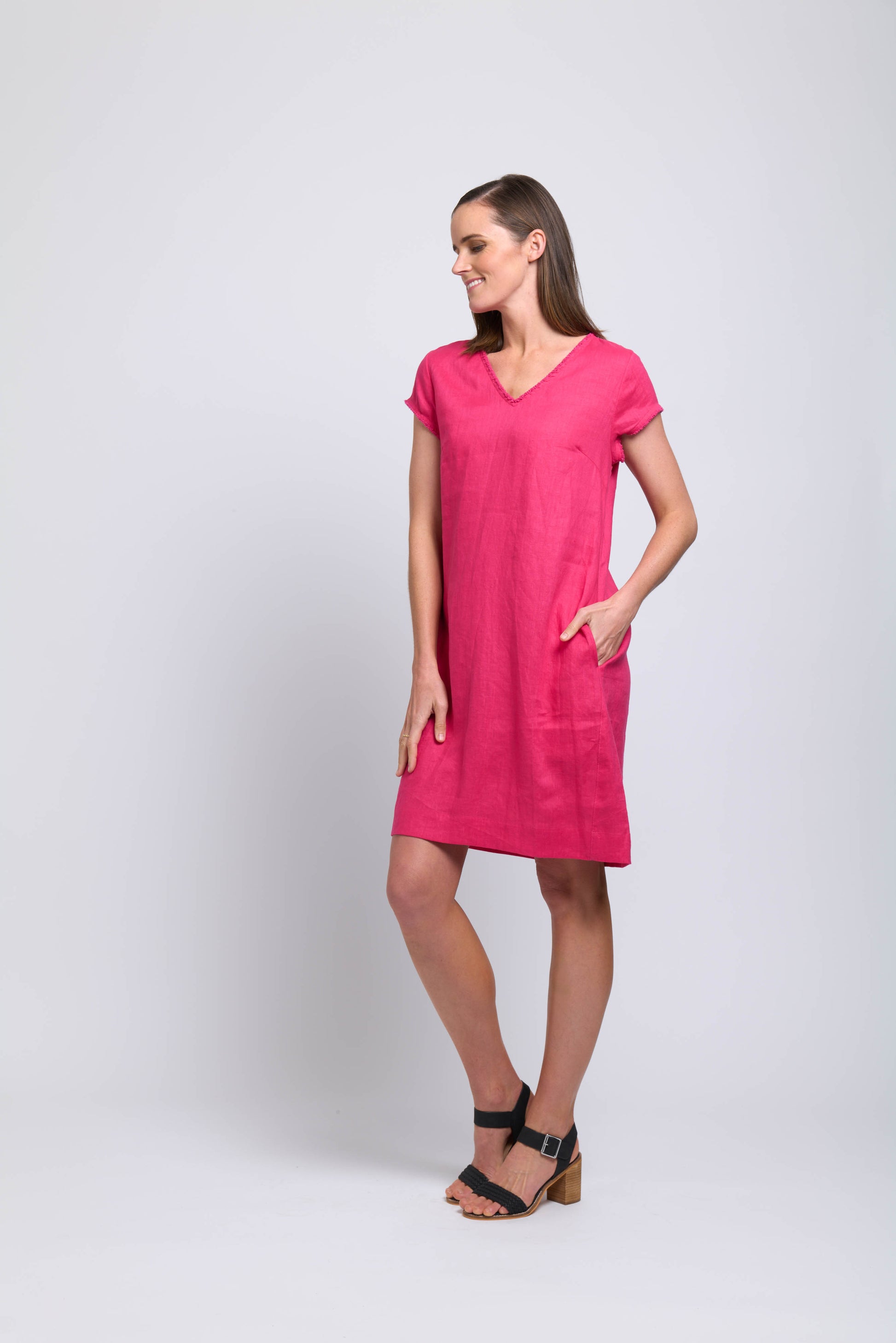 A woman stands against a plain light background, smiling in the FOIL Fringe Festival Dress FO7907 Pink Flash—a bright pink, knee-length dress with short sleeves and a V-neckline. She has her left hand in her pocket and looks to the side wearing black heeled sandals.