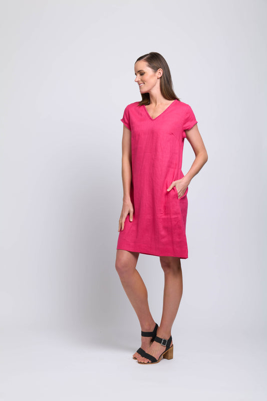 A woman stands against a plain light background, smiling in the FOIL Fringe Festival Dress FO7907 Pink Flash—a bright pink, knee-length dress with short sleeves and a V-neckline. She has her left hand in her pocket and looks to the side wearing black heeled sandals.