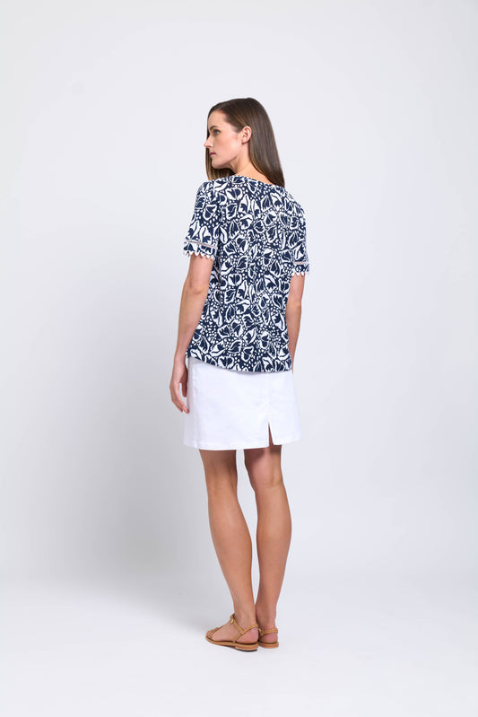 A woman faces away, wearing FOIL’s All the Trimmings Linen Top (CFO7826)—a navy and white patterned short-sleeve top—paired with a white linen knee-length skirt and brown sandals, styled effortlessly against a plain light backdrop.