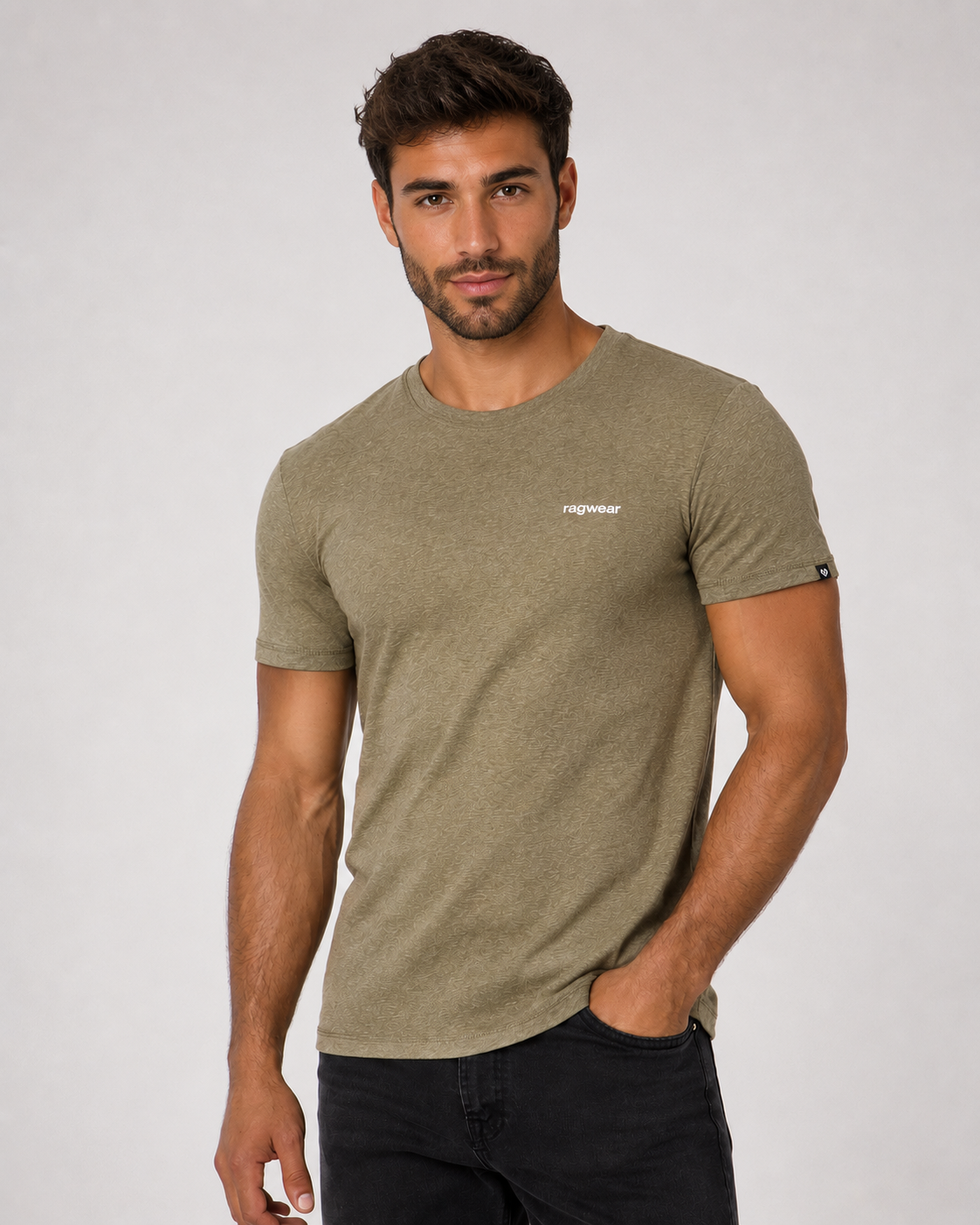 A man with short brown hair and a beard wears the RAGWEAR Hallys Tee 2612-15004 in Dark Olive, featuring a small white chest logo, paired with black jeans. He stands against a light background, one hand in his pocket.