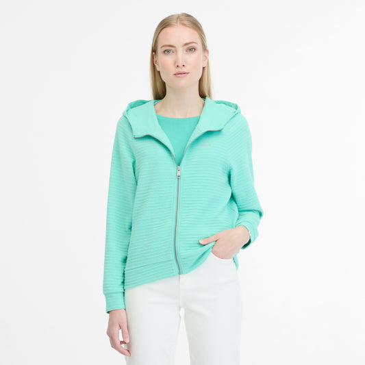 A person stands against a white background, wearing white pants and the TERYA CREPE 2511-30031-5002 by RAGWEAR—a mint green zip-up hooded jacket with a ribbed design. They look ahead neutrally, with one hand in their pocket, exuding a laid-back vibe.