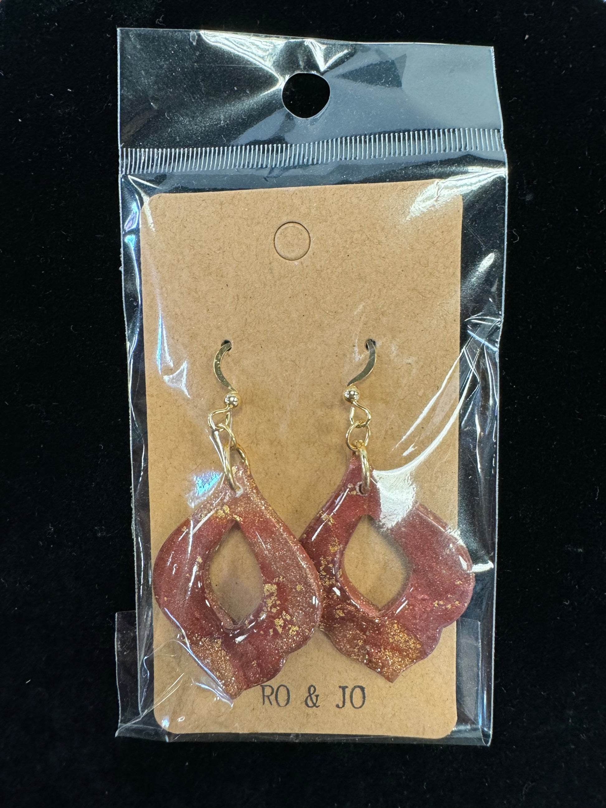 A pair of handmade Ro&Jo Earrings 430 in a clear plastic bag with a cardboard backing. These red resin earrings feature gold accents and have a unique, abstract shape. The packaging is labeled "RO&JO" at the bottom, highlighting their artisanal craftsmanship.