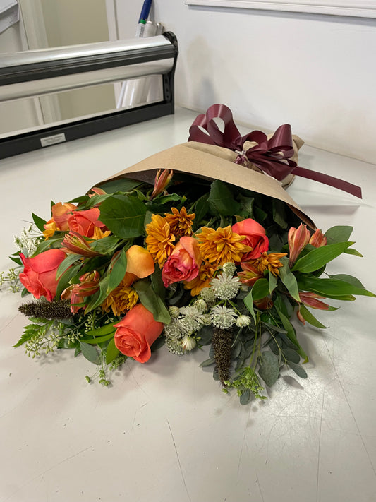 A stunning bouquet called Designer’s Choice Freshcuts from Posies Flowers & Fashion is resting on a white surface. The arrangement features vibrant orange and pink roses, cheerful yellow chrysanthemums, elegant white flowers, and assorted green foliage. It is partially wrapped in brown paper and elegantly tied with a burgundy ribbon.