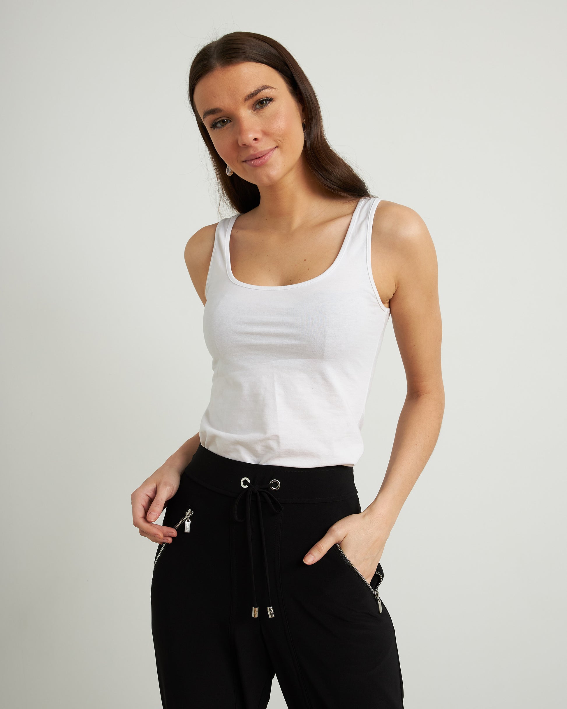 An individual wearing Joseph Ribkoff's Drawstring Waist Pants Style 211317, featuring zipper detailing in black, paired with a white top and black heels. The person stands on a wooden floor against a plain, light-colored background, seamlessly combining comfort and style.