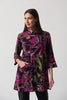 A woman with long brown hair is wearing the Joseph Ribkoff Black/Multi Printed Cowl Neck Tunic Style 233056, featuring an abstract pattern in black, pink, and green hues. She stands against a plain white background, with one hand resting in her pocket and a neutral expression on her face.