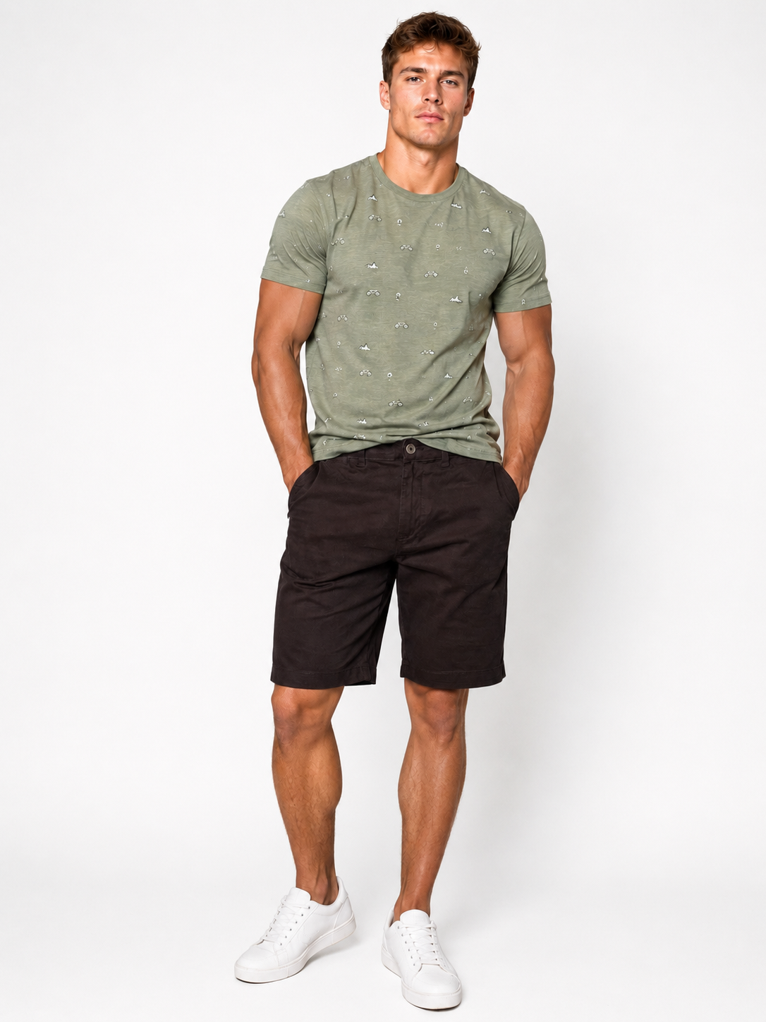 A man stands against a white background, wearing a light green patterned t-shirt, Silver Jeans Co. Ashton Stretch Twill Walking Shorts in black, and white sneakers. His hands are in his shorts pockets and he looks directly at the camera.