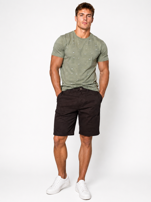 A man stands against a white background, wearing a light green patterned t-shirt, Silver Jeans Co. Ashton Stretch Twill Walking Shorts in black, and white sneakers. His hands are in his shorts pockets and he looks directly at the camera.