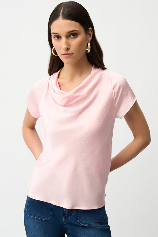 A woman with long brown hair and hoop earrings wears the Joseph Ribkoff Recycled Satin Top With Cowl Neckline 261195 in pale pink, standing against a plain white background with her hands behind her back.