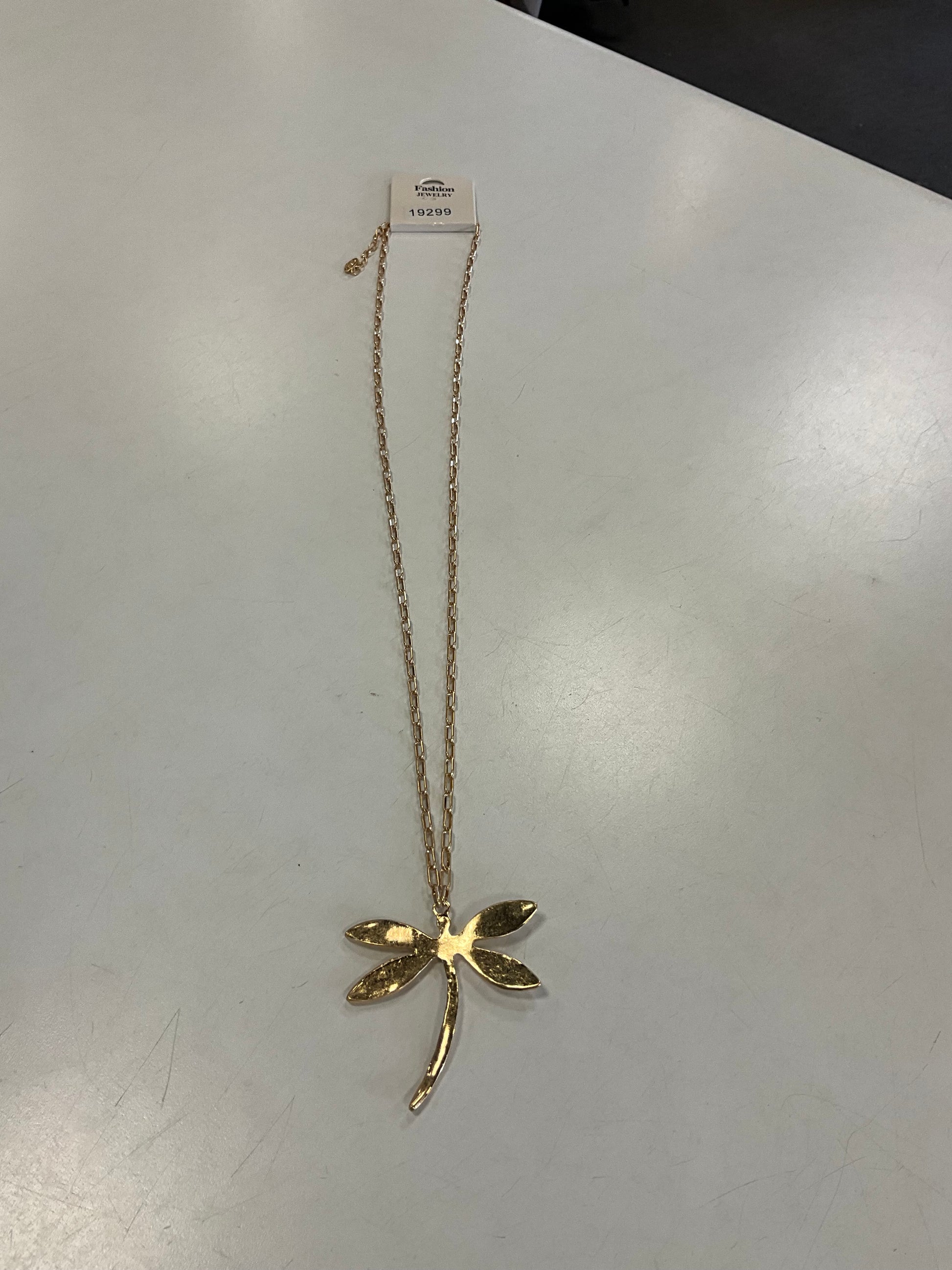 The Ava 22 Gold Necklace from Elise Accessories, featuring a hammered dragonfly-shaped pendant, is displayed on a white surface with a price tag attached to the chain near the top.