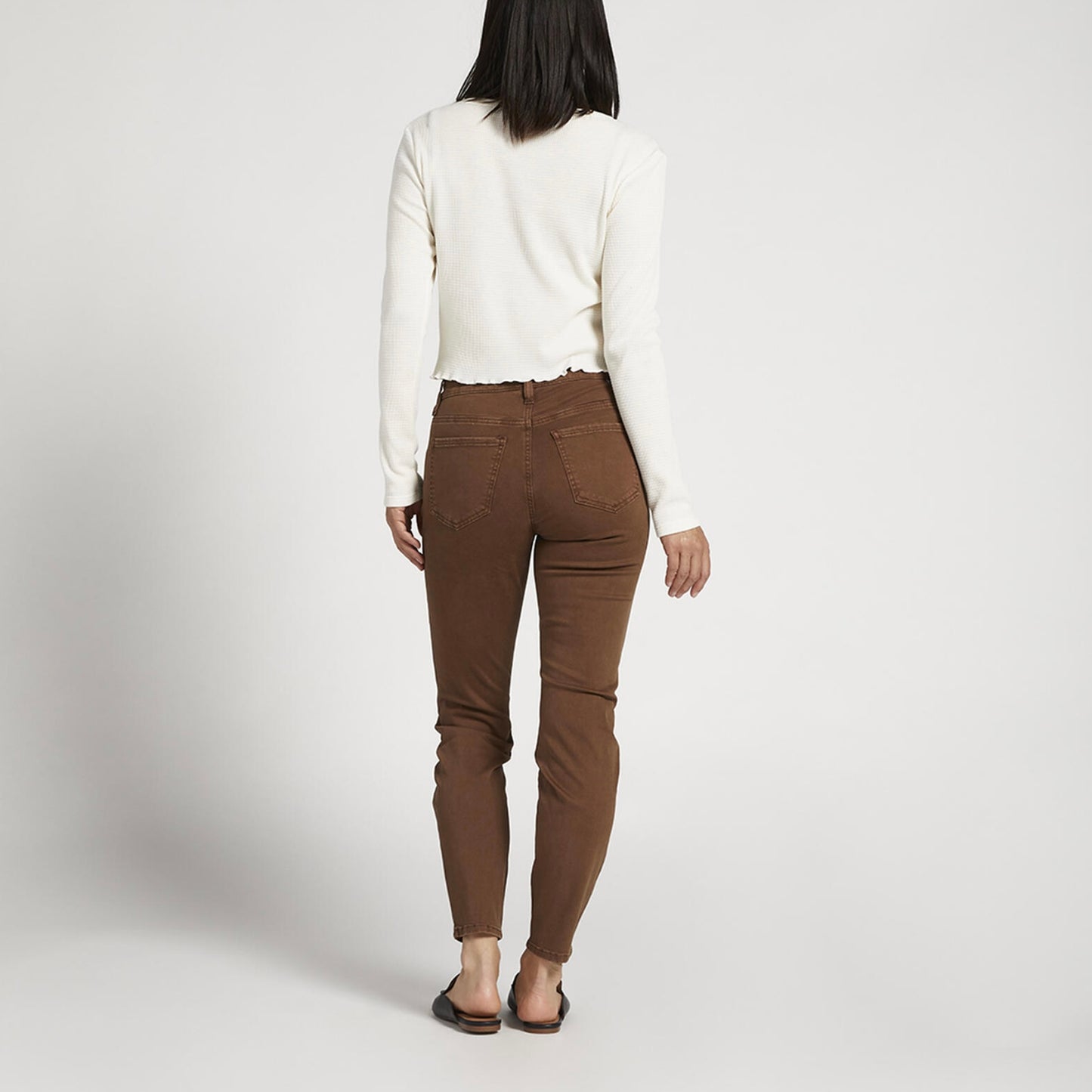 A person wearing a long-sleeved white shirt, Cecilia Mid Rise Skinny Pants - JAG in brown, and black slip-on shoes stands against a plain white background. They are slightly turned to the right and have one arm resting casually by their side.