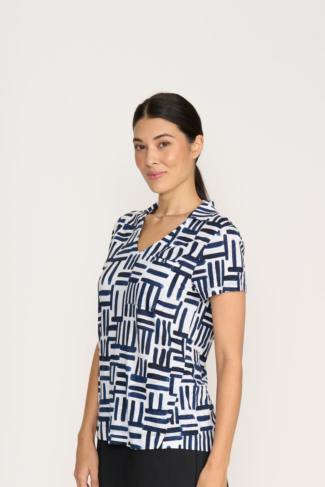 A woman with dark hair pulled back wears the FOIL Team Player Top FO8247 Block & Roll, a short-sleeve V-neck knit linen top featuring a navy and white geometric pattern, standing against a plain light background and smiling slightly.