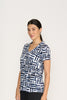 A woman with dark hair pulled back wears the FOIL Team Player Top FO8247 Block & Roll, a short-sleeve V-neck knit linen top featuring a navy and white geometric pattern, standing against a plain light background and smiling slightly.