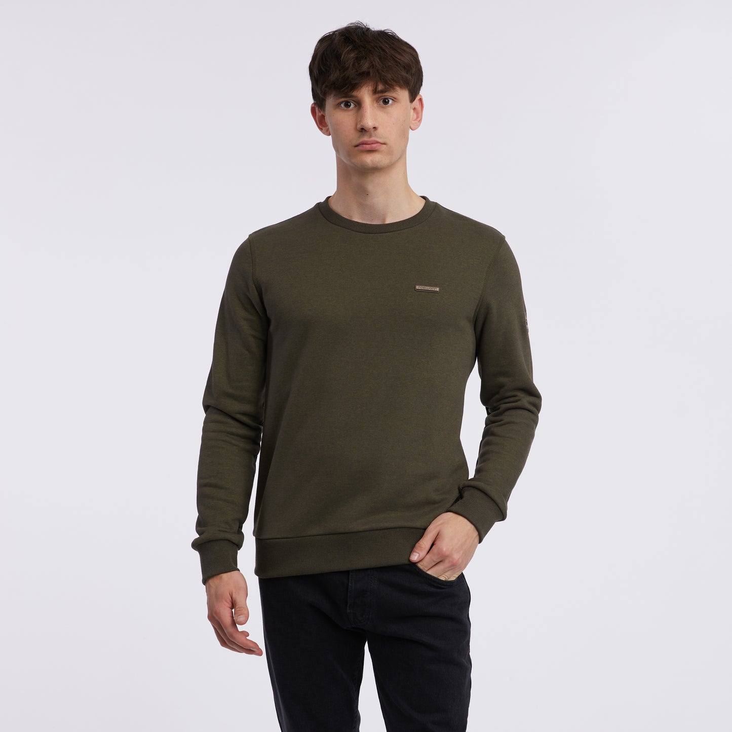 A person with short brown hair is standing in front of a white background. They are wearing the dark green long-sleeved Inddie Core Sweatshirt 9002-30001-5010 by RAGWEAR and black pants. Their right hand is slightly extended while their left hand is relaxed by their side, showcasing the casual elegance of streetwear jackets crafted from vegan materials.