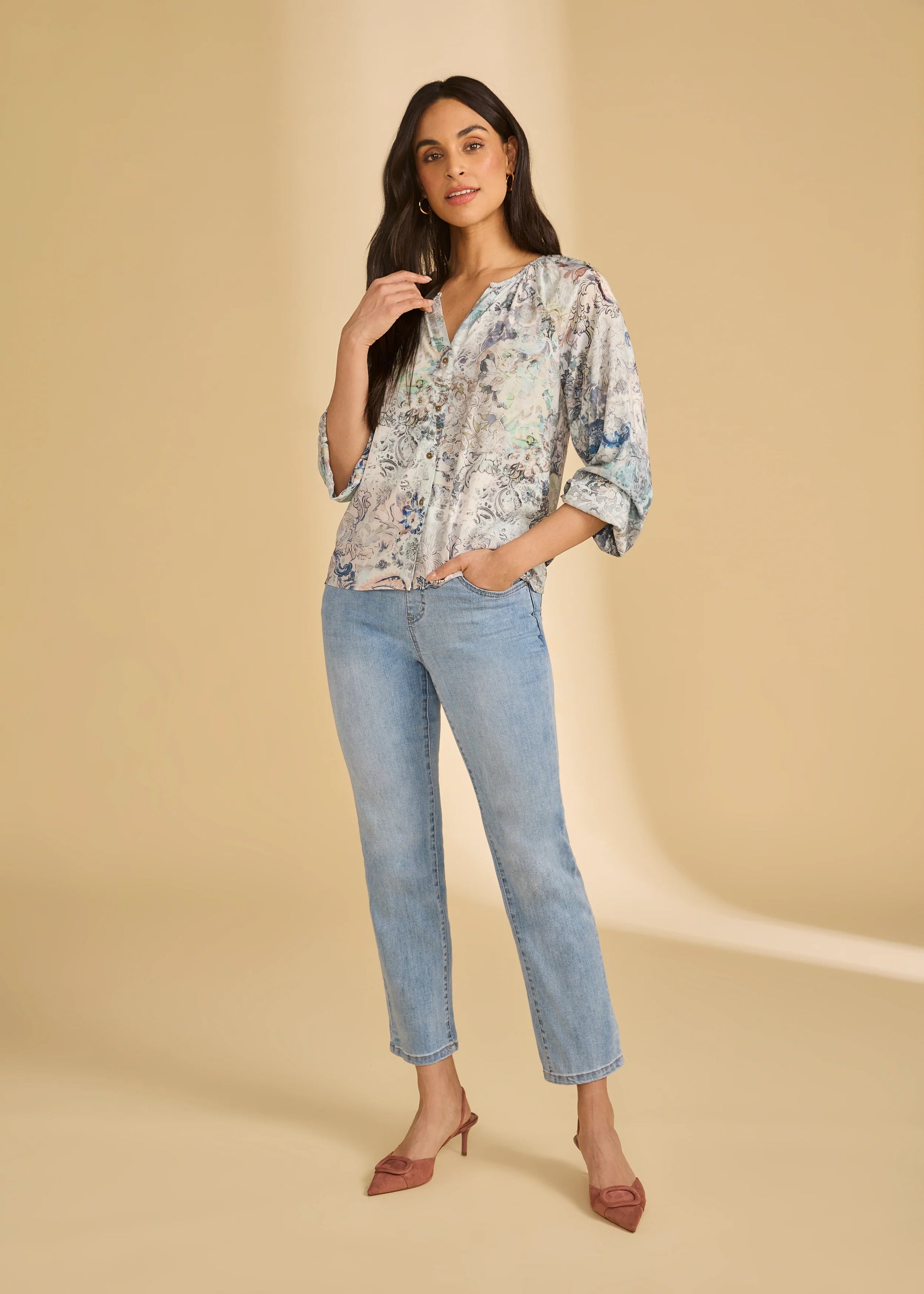 A woman stands against a beige background in French Dressing Jeans' Basic Pull-on Straight Ankle Denim 2442669 in Light Wash, styled with a loose floral blouse, mauve heels, and one hand in her pocket. She has long hair.