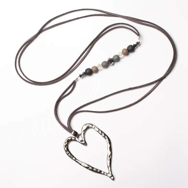 The Suzie Blue Long Open Heart Necklace with Offset Beads in GREY features a large, hammered metal heart pendant on a dark brown suede cord. The cord is adorned with small round beads in shades of black, gray, and beige, interspersed with metallic spacer beads. This handmade necklace is elegantly displayed against a white background.